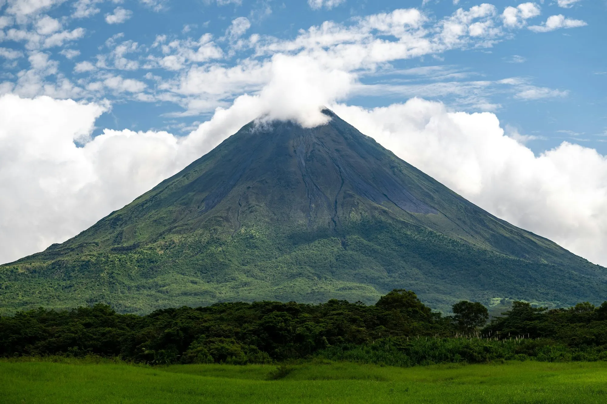 Arenal Volcano with a cloud at its summit above green fields in San Carlos